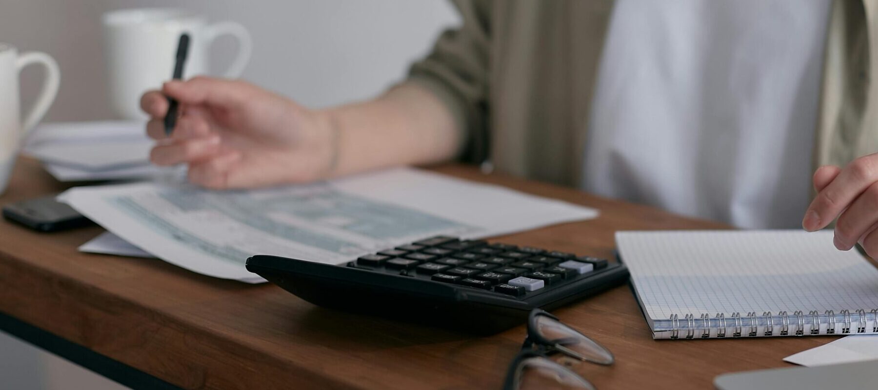 A woman manages finances at home, using a laptop and calculator on a wooden desk.